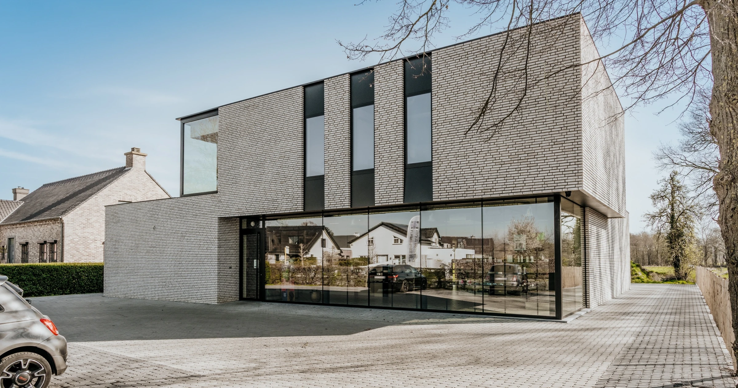 Een modern, strak vormgegeven kantoorpand of woning met een opvallende gevel van lichtgrijze bakstenen en grote glaspartijen op de begane grond. De bovenverdieping heeft verticale ramen gevat in zwarte panelen. De voorzijde is voorzien van een geasfalteerd terrein met geparkeerde auto's en een groot raam.