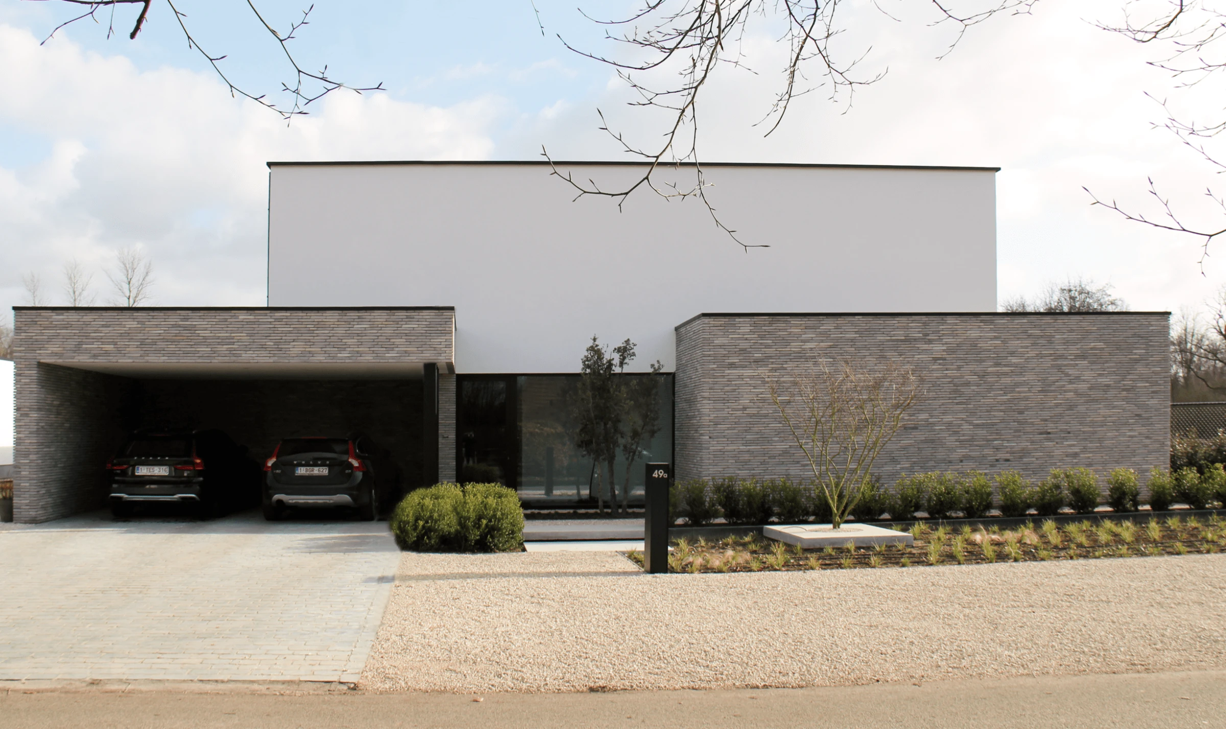 Vooraanzicht van een moderne, strakke woning met een witte bovenverdieping, een met grijze steen beklede begane grond en een open carport met twee auto's.