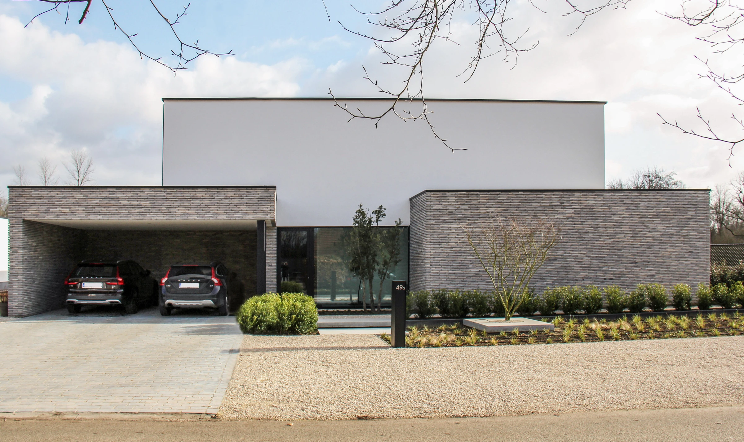 Vooraanzicht van een moderne, strakke woning met een witte bovenverdieping, een met grijze steen beklede begane grond en een open carport met twee auto's.