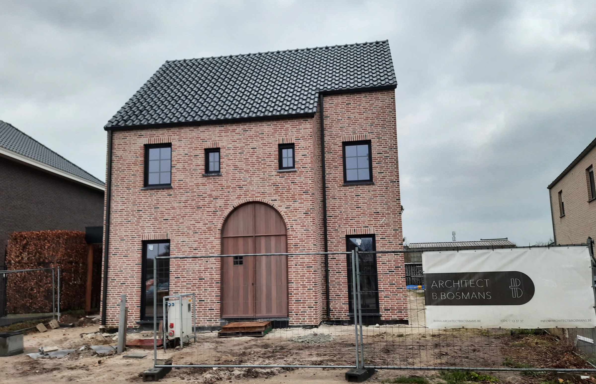 Werffoto van een modern landelijke woning in gemêleerde rode baksteen met een donker zadeldak en een opvallende houten boogdeur.