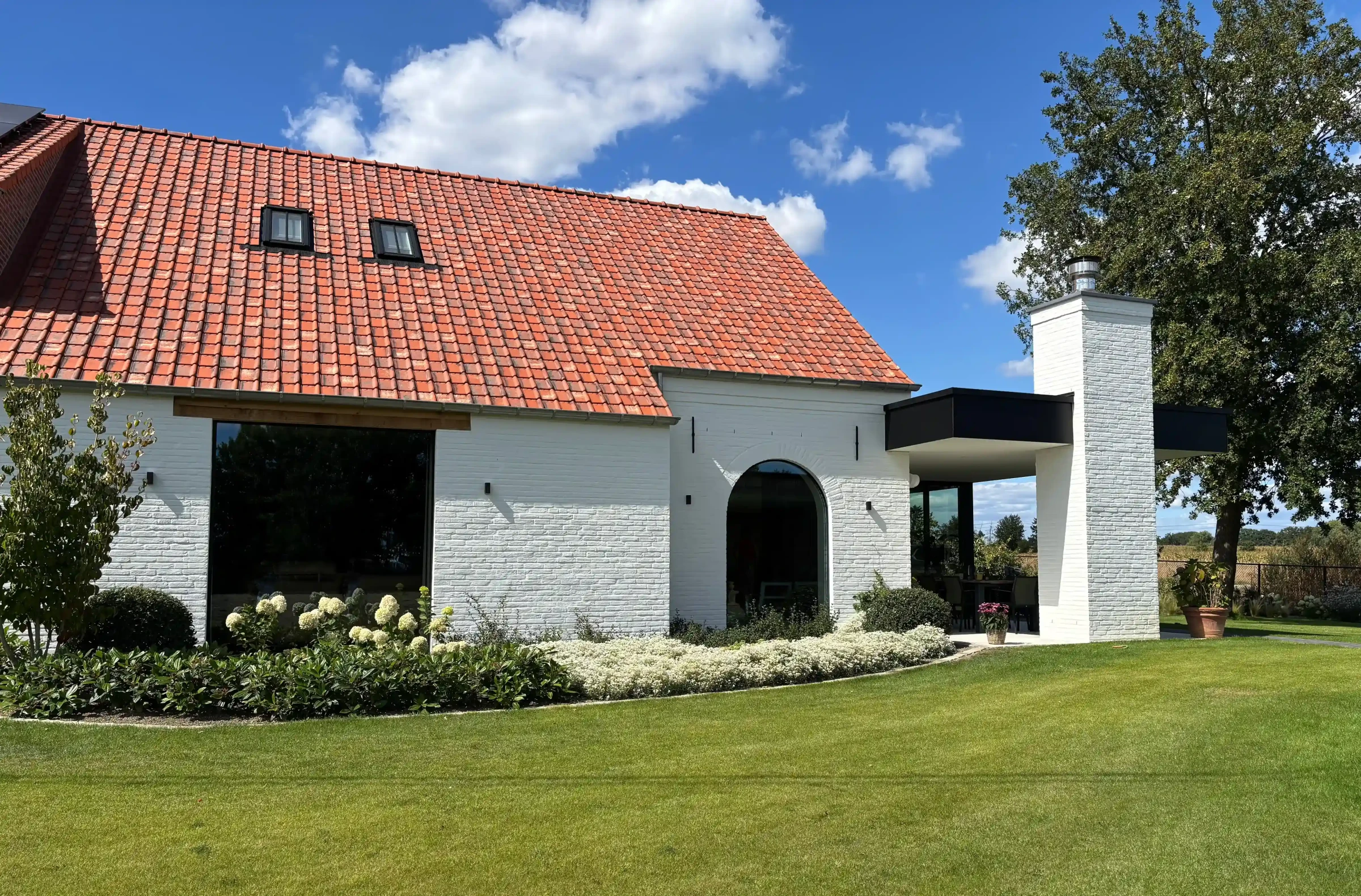 Een modern gerenoveerde hoeve met witgekalkte bakstenen gevels, een oranje pannendak en een strakke zwarte aanbouw met een overdekt terras, omgeven door een verzorgde tuin met gazon en witte hortensia's.