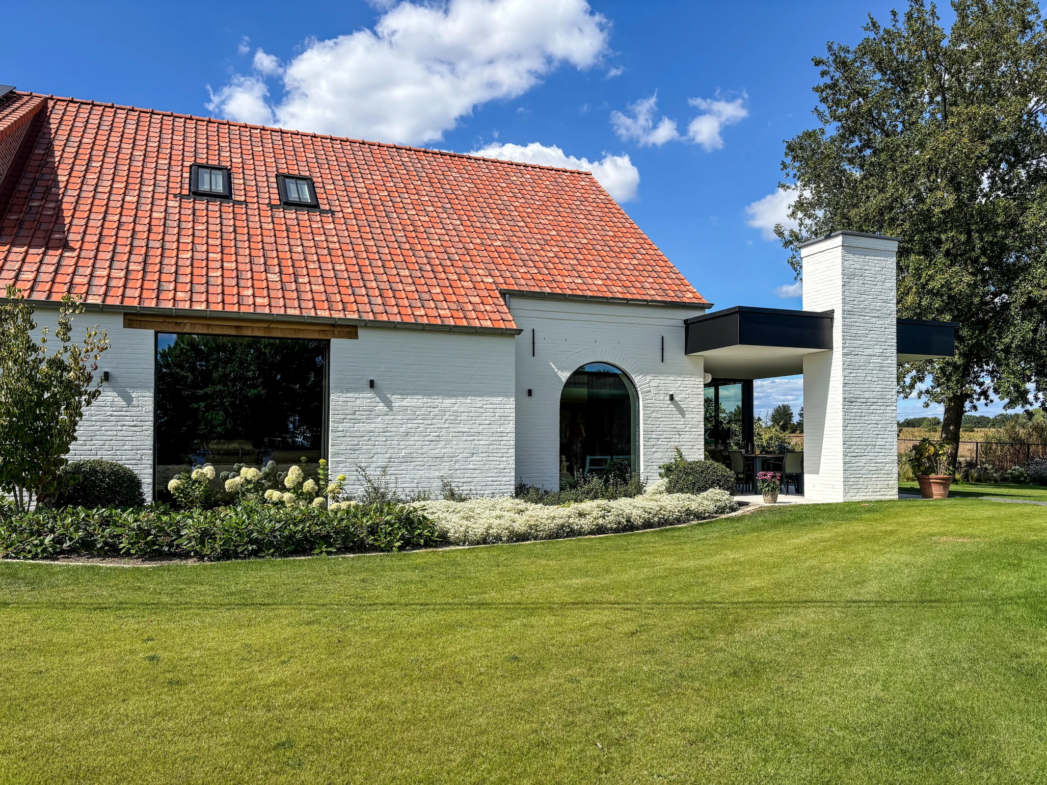 Een modern gerenoveerde hoeve met witgekalkte bakstenen gevels, een oranje pannendak en een strakke zwarte aanbouw met een overdekt terras, omgeven door een tuin met gazon en witte hortensia's.